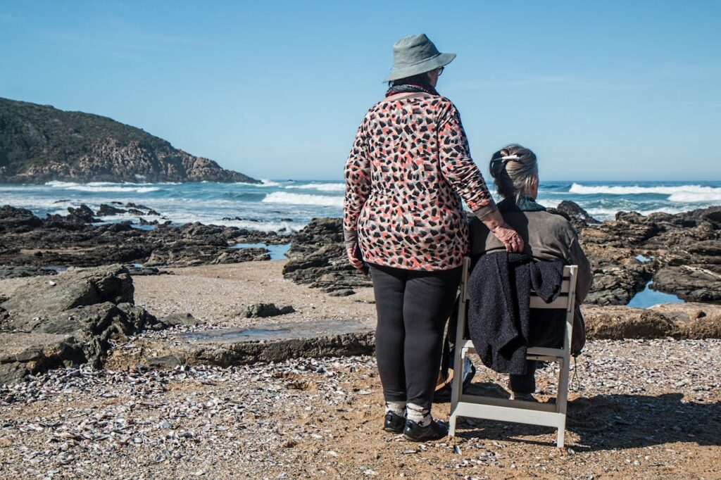 women friends friendship helping together 160767 Two senior women enjoying a peaceful moment by the rocky beach, embracing friendship and serenity.